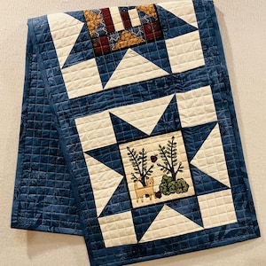 May include: A quilted table runner with a blue background and star-shaped designs. The stars are cream-colored with a central square featuring a cat, trees, and apples. The runner is approximately 60 inches long.