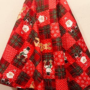 May include: Red Christmas tree skirt with a patchwork design. The skirt features squares of red plaid, red with white polka dots, and red with white snowflakes. Embroidered images of Santa Claus, snowmen, and reindeer are scattered across the skirt.