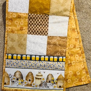 May include: A quilted table runner with a patchwork design. The top features squares of various fabrics in shades of yellow, white, and brown. The bottom section has a bee and beehive pattern. The edges are finished with a floral print.