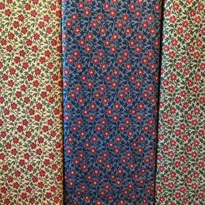 Könnte beinhalten: Drei Stoffmuster mit einem sich wiederholenden Blumenmuster. Der Stoff links ist beige mit grünen Blättern und roten Blüten. Der mittlere Stoff ist blau mit grünen Blättern und roten Blüten. Der Stoff rechts ist grün mit grünen Blättern und roten Blüten.