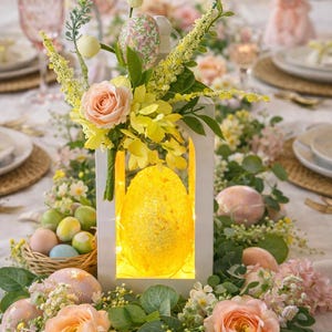 May include: A festive Easter table setting with a white lantern centerpiece, illuminated with a glowing egg. The table is adorned with pastel-colored eggs, fresh flowers, and a decorative bunny, creating a spring-themed ambiance.
