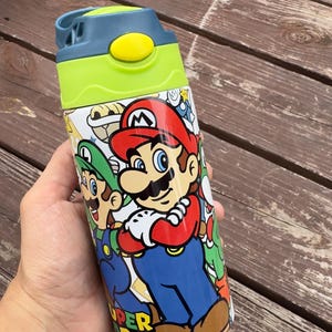 Puede incluir: Una botella de agua colorida con los personajes Mario y Luigi de la serie de videojuegos Super Mario Bros. La botella tiene una tapa verde y azul y un cuerpo blanco con las imágenes de los personajes. Las palabras "SUPER MARIO" son visibles.
