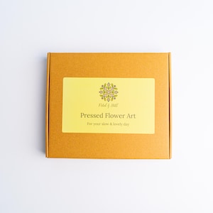 May include: A square, mustard-colored box with a rectangular yellow label. The label reads "Pressed Flower Art" with the text "For your slow & lovely day" below. The box has a floral design in the upper center.