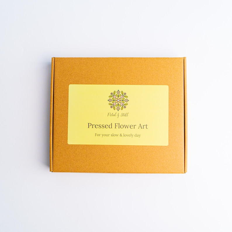 May include: A square, mustard-colored box with a rectangular yellow label. The label reads "Pressed Flower Art" with the text "For your slow & lovely day" below. The box has a floral design in the upper center.