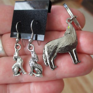 May include: A pair of silver-toned metal earrings featuring a howling wolf design attached to a black display card, alongside a matching silver-toned metal keychain featuring a howling wolf with a small yellow accent on the eye area.