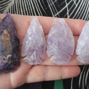 May include: Four amethyst arrowheads in varying shades of purple are displayed. The arrowheads are hand-carved and have a textured surface. The amethyst pieces are held in a hand, showcasing their natural beauty and unique shapes.