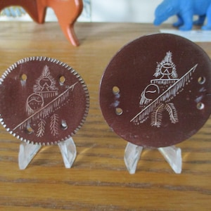 May include: Two brown, circular decorative pieces with white etched designs of a figure with a feathered headdress and a spear. One is round with a scalloped edge, the other is smooth. Both are displayed on clear plastic stands.