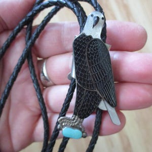 May include: A bolo tie featuring a silver and black eagle pendant with a small, oval turquoise stone accent at the bottom. The eagle is attached to a black braided leather cord with silver-toned metal tips at the ends of the cord strands.