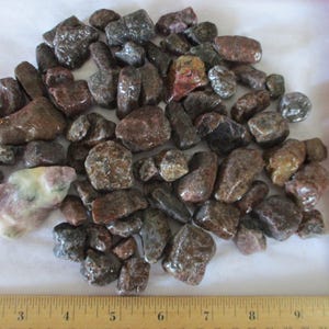 May include: A collection of polished, tumbled stones in shades of brown, gray, and reddish-brown. The stones vary in size and shape, with a few lighter-colored stones mixed in. A wooden ruler is visible along the right side and bottom.