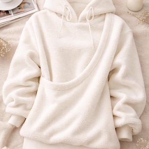 May include: Cream-colored, plush hoodie with a unique asymmetrical design. Features a hood with drawstrings, long sleeves, and a kangaroo pocket. The soft, textured fabric suggests a cozy, comfortable feel. A stylish and warm addition to any wardrobe.