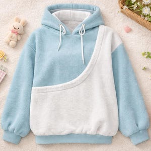 May include: A light blue and white fleece hoodie with a kangaroo pocket. The hoodie has a drawstring hood and long sleeves. A plush bunny, a strawberry-shaped pillow, and a small basket of flowers are also in the image.