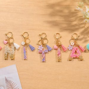 May include: A collection of personalized keychains featuring resin letters filled with dried flowers. Each keychain has a gold-colored clasp, tassel, and charm, with a heart or flower accent. The letters are in various colors, including pink, purple, and blue.