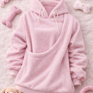 May include: A light pink fleece hoodie with a kangaroo pouch, white drawstrings, and ribbed cuffs. The image also includes pink dog toys, such as a bone-shaped toy, a ring, and a plush dog toy.