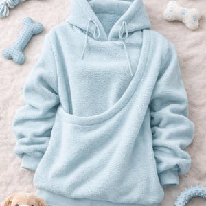 May include: A light blue pet carrier hoodie with a kangaroo pouch, surrounded by dog toys. The hoodie has a drawstring hood and long sleeves. The toys include a rope toy, bone-shaped plush toys, and a plush dog toy.
