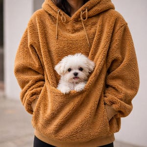May include: A brown, hooded sweatshirt with a kangaroo pocket, holding a small, fluffy white dog. The sweatshirt has a soft, textured appearance and a drawstring hood. The dog is a Maltese, with its tongue slightly out.