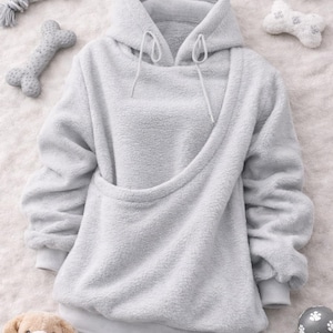 May include: A light gray, plush hoodie with a unique sling-style pocket. The hoodie features a drawstring hood and ribbed cuffs. Dog toys, including a bone-shaped toy and a plush dog, are arranged around the hoodie, suggesting a pet-themed product.