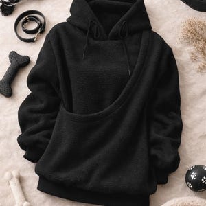 May include: Black hooded sweatshirt with a kangaroo pouch, designed for pet carrying. The image also shows a dog leash, toys, a dog bowl, and a pet bed. The sweatshirt is made of a soft, textured material.