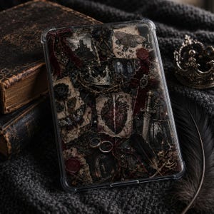 Pode incluir: Uma capa protetora transparente para tablet com uma colagem de estilo gótico escuro. O design inclui imagens de uma coroa, um coração, uma adaga, uma mariposa, uma rosa e um castelo, acentuados com fitas de veludo vermelho, selos de cera e uma pena preta.