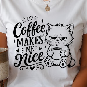 May include: A white short-sleeved t-shirt featuring a black graphic print of a grumpy cat holding a coffee mug. The text reads Coffee MAKES ME Nice, accompanied by small heart and star icons. The design is centered on the front of the shirt.