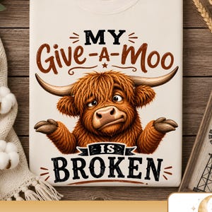 May include: A cream-colored t-shirt featuring a graphic of a brown, shaggy Highland cow with long horns and a shrug gesture. The text reads MY Give-a-Moo IS BROKEN in brown and black lettering. The design is centered on the front of the shirt.