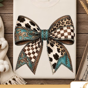 May include: A cream-colored t-shirt featuring a large graphic of a bow. The bow is composed of various patterns including black and white cow print, brown leopard spots, a brown and white checkerboard pattern, and turquoise floral filigree.