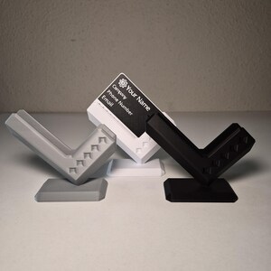 以下が含まれることがあります： グレー、白、黒の3つの名刺入れ。各ホルダーは、ベースを備えたユニークな角度の付いたデザインになっています。白いホルダーには、「Your Name」、「Company」、「Phone Number」、「Email」というテキストが記載された名刺が表示されます。