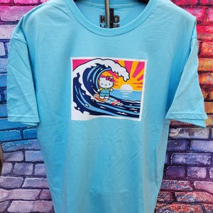 May include: A light blue short-sleeve t-shirt featuring a graphic print of a white cat character with a pink bow surfing on a large blue wave. The background of the graphic shows a yellow sun and pink rays. The shirt has a crew neckline.