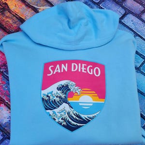 May include: A light blue hooded sweatshirt featuring a graphic on the back. The graphic is a shield-shaped emblem with the text SAN DIEGO in white letters above a stylized ocean wave, a setting sun, and horizontal stripes in pink, orange, and yellow.