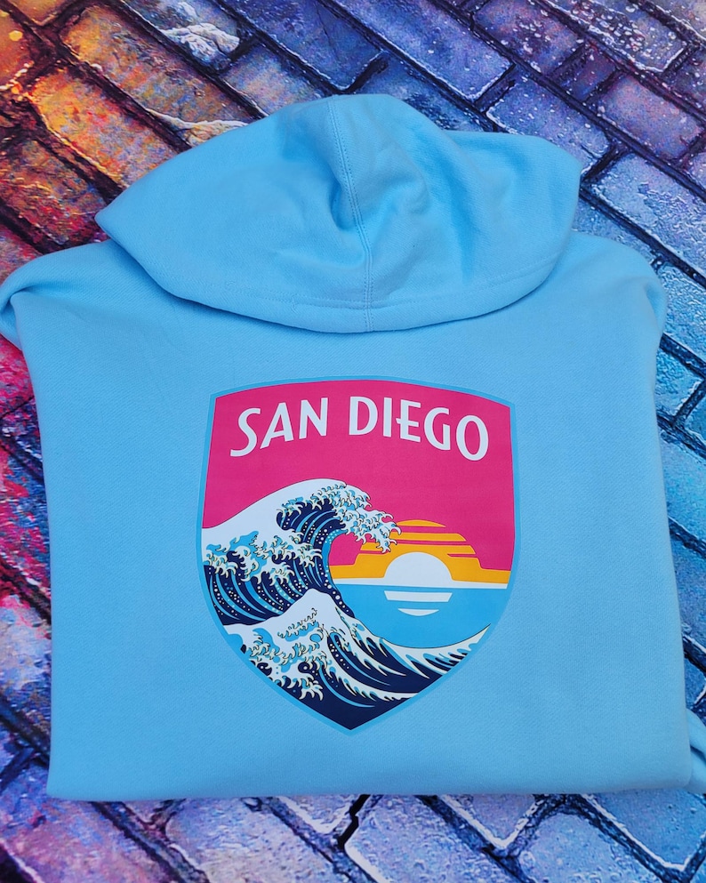 May include: A light blue hooded sweatshirt featuring a graphic on the back. The graphic is a shield-shaped emblem with the text SAN DIEGO in white letters above a stylized ocean wave, a setting sun, and horizontal stripes in pink, orange, and yellow.