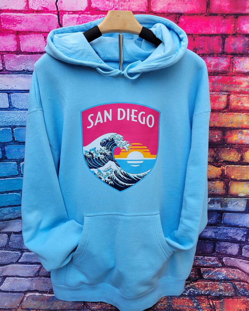 May include: A light blue hooded sweatshirt featuring a central graphic shield. The shield displays the text SAN DIEGO above a stylized ocean wave, a setting sun, and horizontal stripes in shades of pink, orange, yellow, and light blue.