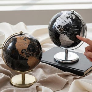 Puede incluir: Dos globos decorativos, uno con base de latón y otro con base plateada, sobre una superficie crema. Los globos son negros con continentes blancos y beige. Un dedo señala el globo de la derecha.