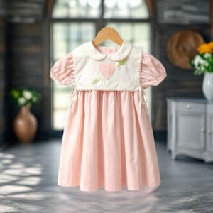 May include: A light pink baby dress with a white bib-style top featuring a pink heart and green leaf design. The dress has short, puffy sleeves and is hanging on a wooden hanger. The dress is made of soft fabric.