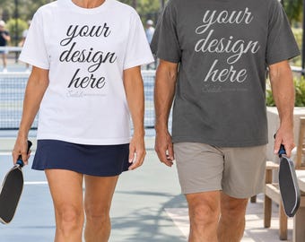 May include: A white short-sleeved t-shirt and a dark gray short-sleeved t-shirt, both featuring the text 'your design here' in a cursive font on the chest. Each shirt is paired with athletic bottoms and held pickleball paddles.