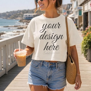 May include: A cream-colored cropped t-shirt featuring the black text "your design here" and "Selah MOCKUP STUDIO" printed on the front. The shirt is paired with light blue denim shorts with a frayed hem and a woven tan shoulder bag.