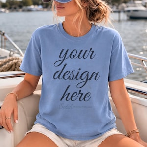 Può includere: Una maglietta azzurra con la scritta "your design here" in corsivo. La maglietta è a maniche corte e ha un girocollo. Il testo "Selah MOCKUP STUDIO" è stampato sotto. La modella indossa la maglietta su una barca.