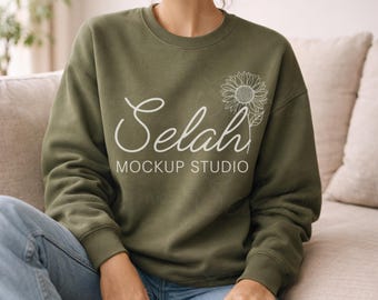 May include: An olive green crew neck sweatshirt featuring the white text "Selah MOCKUP STUDIO" and a white sunflower design. The sweatshirt is being worn and is a casual style.