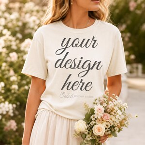 Peut inclure: T-shirt crème avec le texte "your design here" en écriture cursive. Le t-shirt fait partie d'une maquette, et la personne tient un bouquet de fleurs. Le texte "Selah Mockup Studio" est en bas.