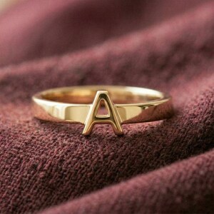 May include: A gold-colored ring with the letter "A" as the central design element. The ring is set against a textured, maroon fabric background, creating a contrast between the shiny metal and the matte textile.