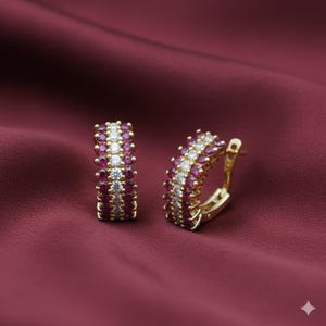 Pode incluir: Um par de brincos de argola dourados. Cada brinco é adornado com fileiras alternadas de pequenas pedras preciosas redondas, vermelhas e brancas. Os brincos são colocados sobre um fundo vermelho escuro, criando um contraste marcante.