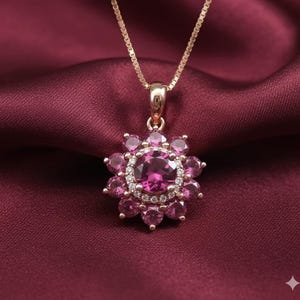 May include: A gold-colored necklace with a pendant featuring a large, round, pink gemstone surrounded by smaller pink gemstones and a halo of clear crystals. The necklace is set against a deep red fabric.