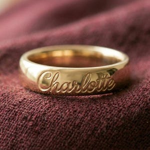 May include: A gold ring engraved with the name "Charlotte" in a cursive script. The ring sits on a maroon fabric. This personalised ring is a piece of jewellery.
