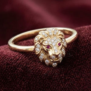 Könnte beinhalten: Ein goldener Ring mit einem Löwenkopf-Design, besetzt mit Diamanten und Rubinaugen. Der Ring liegt auf einem dunkelroten Samthintergrund, der die feinen Details und die Handwerkskunst hervorhebt.