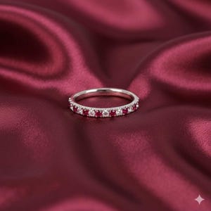 Puede incluir: Un anillo de plata con piedras preciosas rojas y claras alternadas. El anillo se encuentra sobre un fondo de satén rojo oscuro, creando un contraste que resalta el brillo del anillo. Las piedras están espaciadas uniformemente.
