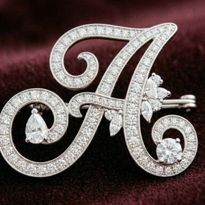 May include: A sparkling, silver-toned brooch in the shape of the letter "A", encrusted with numerous small, clear gemstones. The brooch features a pin closure on the back and is set against a dark burgundy background. The design includes a pear-shaped stone.