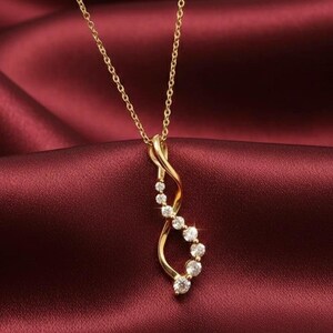 Pode incluir: Um colar com pingente dourado com uma corrente delicada. O pingente apresenta um design curvo e fluido adornado com várias pedras preciosas redondas e transparentes brilhantes. O colar repousa sobre um tecido vermelho escuro semelhante ao cetim.