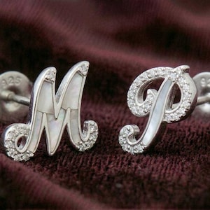 Könnte beinhalten: Ein Paar Ohrstecker aus Sterlingsilber mit den Buchstaben "M" und "P". Jeder Buchstabe ist mit kleinen, funkelnden, klaren Steinen und einer Perlmutt-Einlage besetzt. Die Ohrringe sind vor einem dunkelburgunderfarbenen Hintergrund platziert.