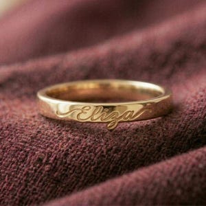 May include: A gold ring with the name "Eliza" engraved in a cursive font. The ring is set against a textured, maroon fabric background. The ring is a simple band design.