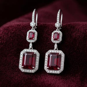 Puede incluir: Pendientes colgantes elegantes con piedras preciosas rectangulares de color rojo intenso engastadas en metal plateado. Las piedras preciosas están rodeadas por un halo de pequeñas piedras blancas brillantes. Los pendientes cuelgan de un poste.
