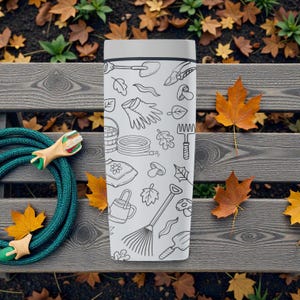 Puede incluir: Un termo blanco con tapa gris, decorado con dibujos lineales negros de herramientas y elementos de jardiner&iacute;a. El termo est&aacute; sobre un banco de madera con una manguera verde y hojas de oto&ntilde;o.