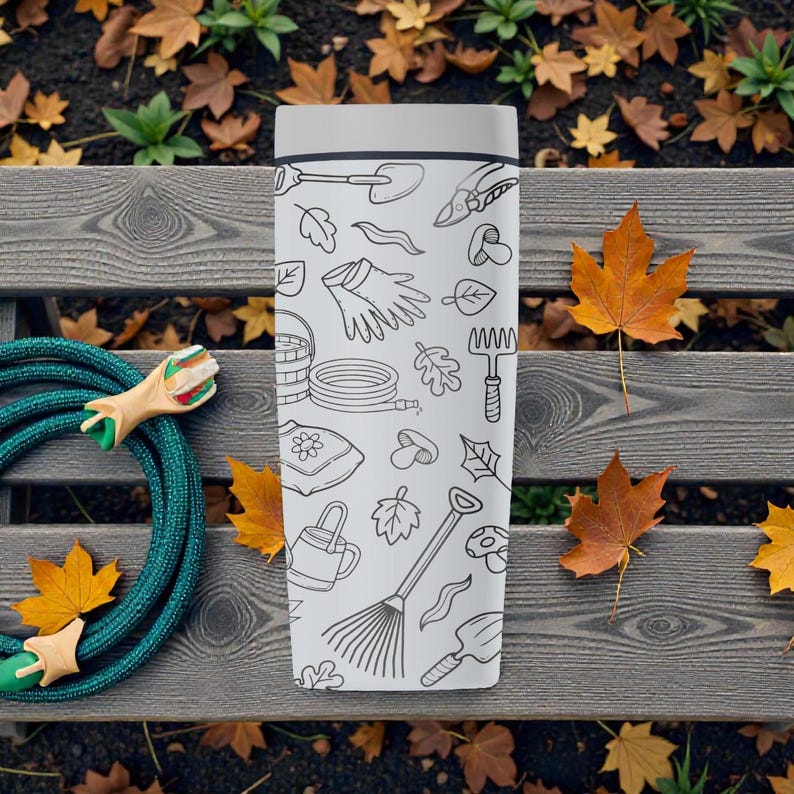 Puede incluir: Un termo blanco con tapa gris, decorado con dibujos lineales negros de herramientas y elementos de jardiner&iacute;a. El termo est&aacute; sobre un banco de madera con una manguera verde y hojas de oto&ntilde;o.